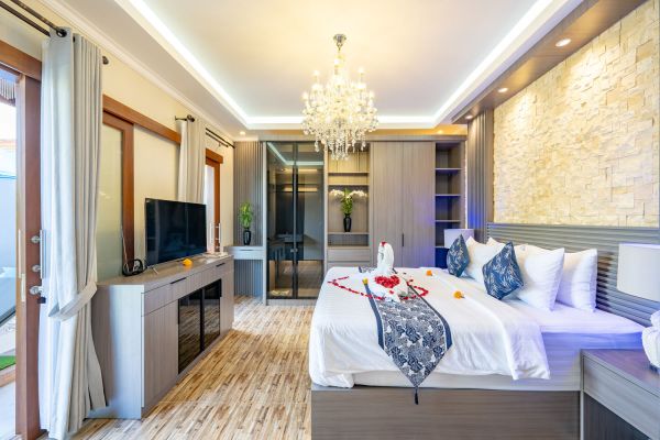 Luxurious Balinese-style hotel bedroom with king-size bed scattered with rose petals, wooden floors, chandelier lighting, and stone accent wall.